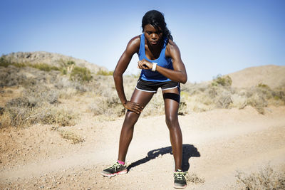 Sporty woman checking time while standing on dirt road against clear sky