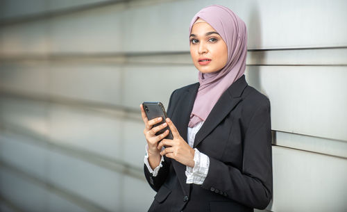 Young woman using phone while standing against wall