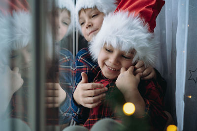 Cute children wearing santa hats and plaid shirts sitting by the window waitig for christmas