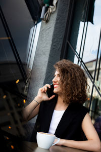 Young pretty curly haired business woman talking by smart phone in cafe. social media concept