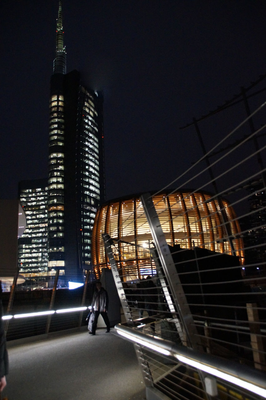 REAR VIEW OF WOMAN WALKING AGAINST ILLUMINATED MODERN BUILDINGS