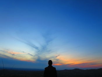 Rear view of silhouette man standing at beach against blue sky during sunset