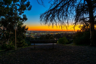 Park bench by trees against sky during sunset