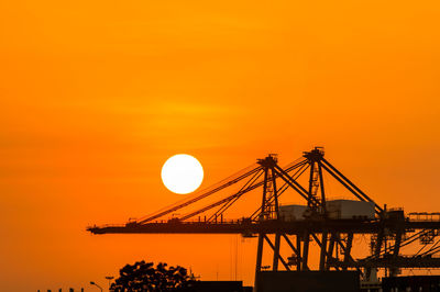 Silhouette cranes at construction site against orange sky