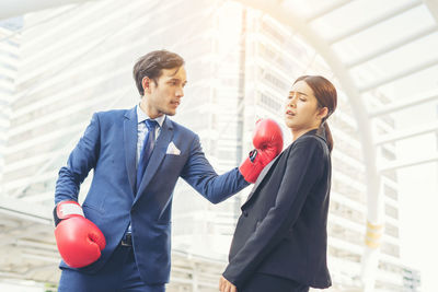 Young businessman hitting colleague with red boxing gloves against buildings in city
