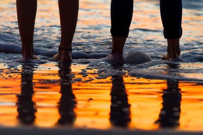 Low section of friends reflecting while standing at beach during sunset