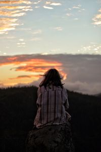 Rear view of woman sitting on mountain against sky during sunset