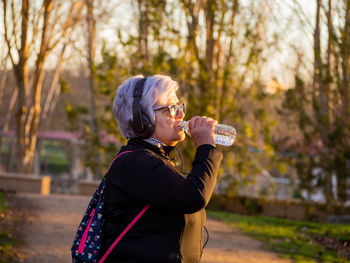 Senior woman drinking water from bottle in park during sunset