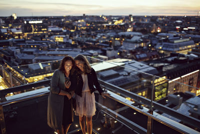 High angle view of smiling female professionals taking selfie while standing on terrace against illuminated cityscape
