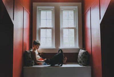 Side view of shirtless cute boy playing with cat while sitting on alcove window seat at home