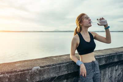 Full length of young woman drinking water at sea against sky