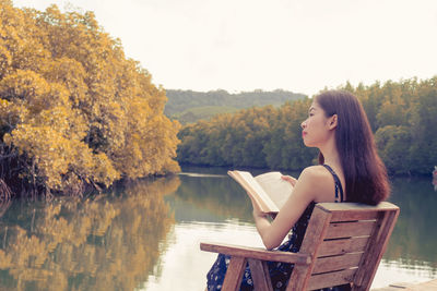 Woman sitting on book by lake against trees