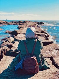 Rear view of woman sitting on rock at beach against sky