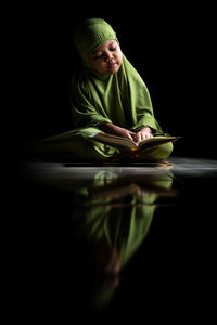 Girl wearing hijab reading koran while sitting on floor against black background