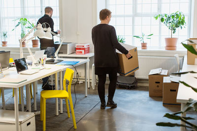 Rear view of businesswoman carrying cardboard box by desk in creative office