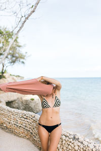 Woman in bikini removing top while standing at beach against sky