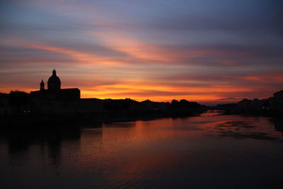 Silhouette buildings by river against sky during sunset