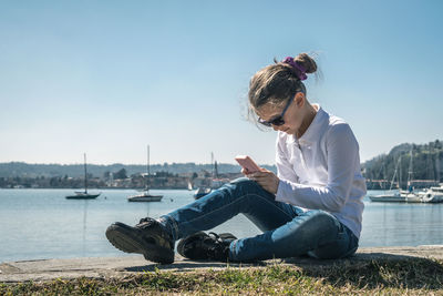 Girl using mobile phone while sitting by sea against sky