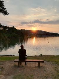 Rear view of man sitting on bench by lake against sky during sunset
