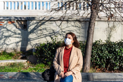 Woman wearing mask looking away sitting against tree