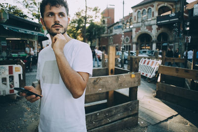 Thoughtful young man holding mobile phone while standing on street in city