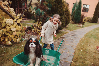 Girl standing by dog in wheelbarrow on footpath