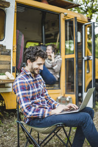 Smiling young man using laptop while sitting on chair against friend in camper vehicle