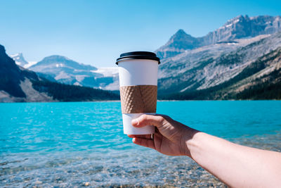 Cropped image of hand holding drink against lake and mountains