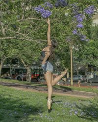 Side view of female ballet dancer jumping for purple flowers on branch at park