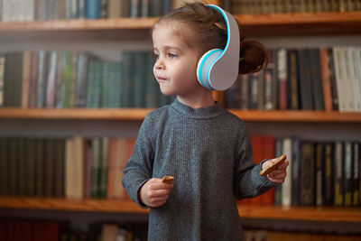 Girl listening music through headphones while standing against bookshelf