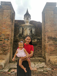 Portrait of woman with baby girl standing at temple against buddha statue 