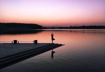 Silhouette woman doing yoga on pier over lake during sunset