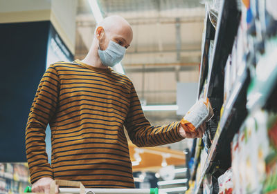 Bald man in medical face mask chooses products in supermarket, concept of coronavirus quarantine