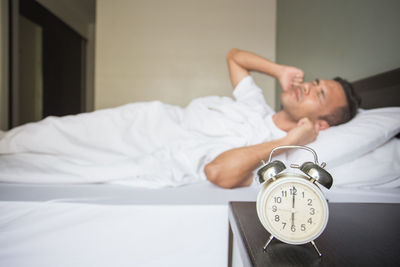 Man with hands covering ears on bed by alarm clock at night table in bedroom