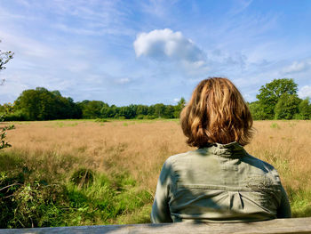 Rear view of woman sitting on a bench, looking over a field under a sky with a small summer cloud