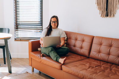 Young smiling woman working on her laptop sitting on the sofa at home