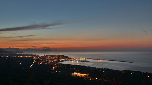 High angle view of illuminated city by sea against sky during sunset