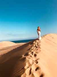 Rear view of woman walking on sand dune in desert against blue sky