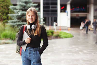 A beautiful fashionable female student in headphones stands in the city with a tablet and a backpack