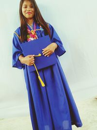 Portrait of young woman in graduation gown standing against wall