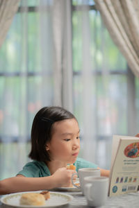 Girl eating food while reading book on table at home