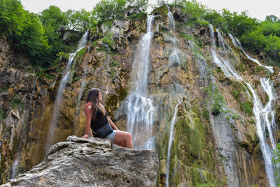 Rear view of woman sitting on rock against waterfall