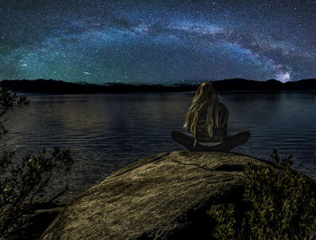 Woman sitting by lake against sky at night