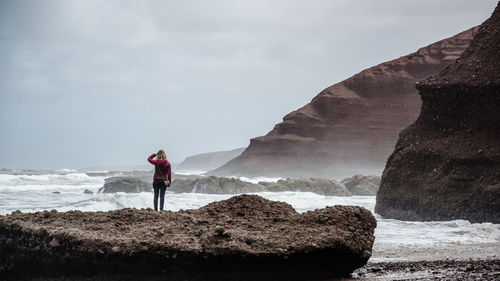 Rear view of woman standing on rock formation by sea against sky