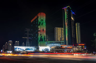 Light trails on road by buildings against sky at night