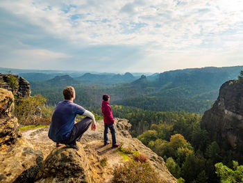 Man hiker sit on edge and woman tourist bellow watching over sunny landscape saxon switzerland.