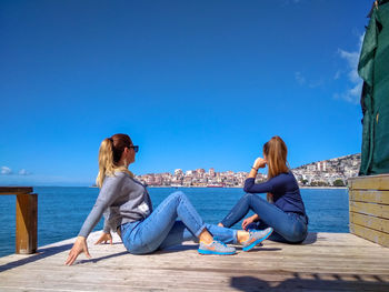 Female friends sitting on pier in sea against blue sky