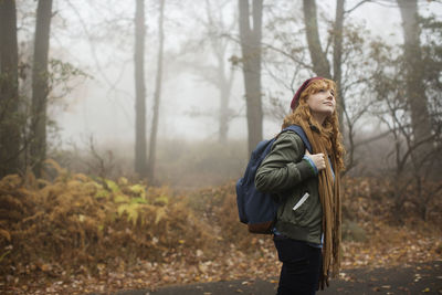 Woman with backpack looking up while standing on road in forest