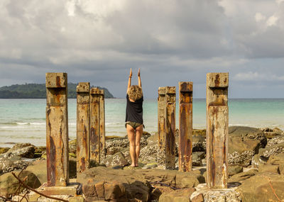 Rear view of woman standing on wooden post in sea against sky