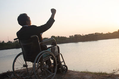 Rear view of man sitting by lake against sky during sunset